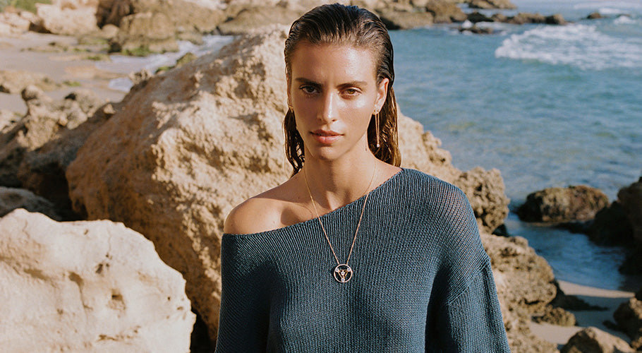 Model standing on rocky shoreline with ocean waves behind, wearing a teal sweater and ANNOORY gold pendant necklace set with lab grown diamonds, embodying elegance and natural energy.